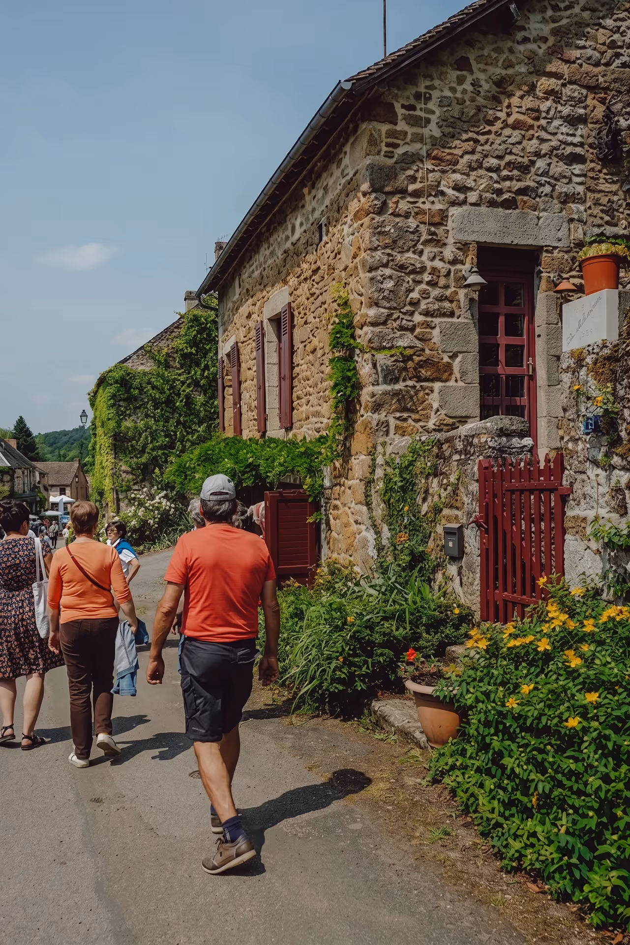 People walking on a street beside a stone house with red shutters and lush green plants.