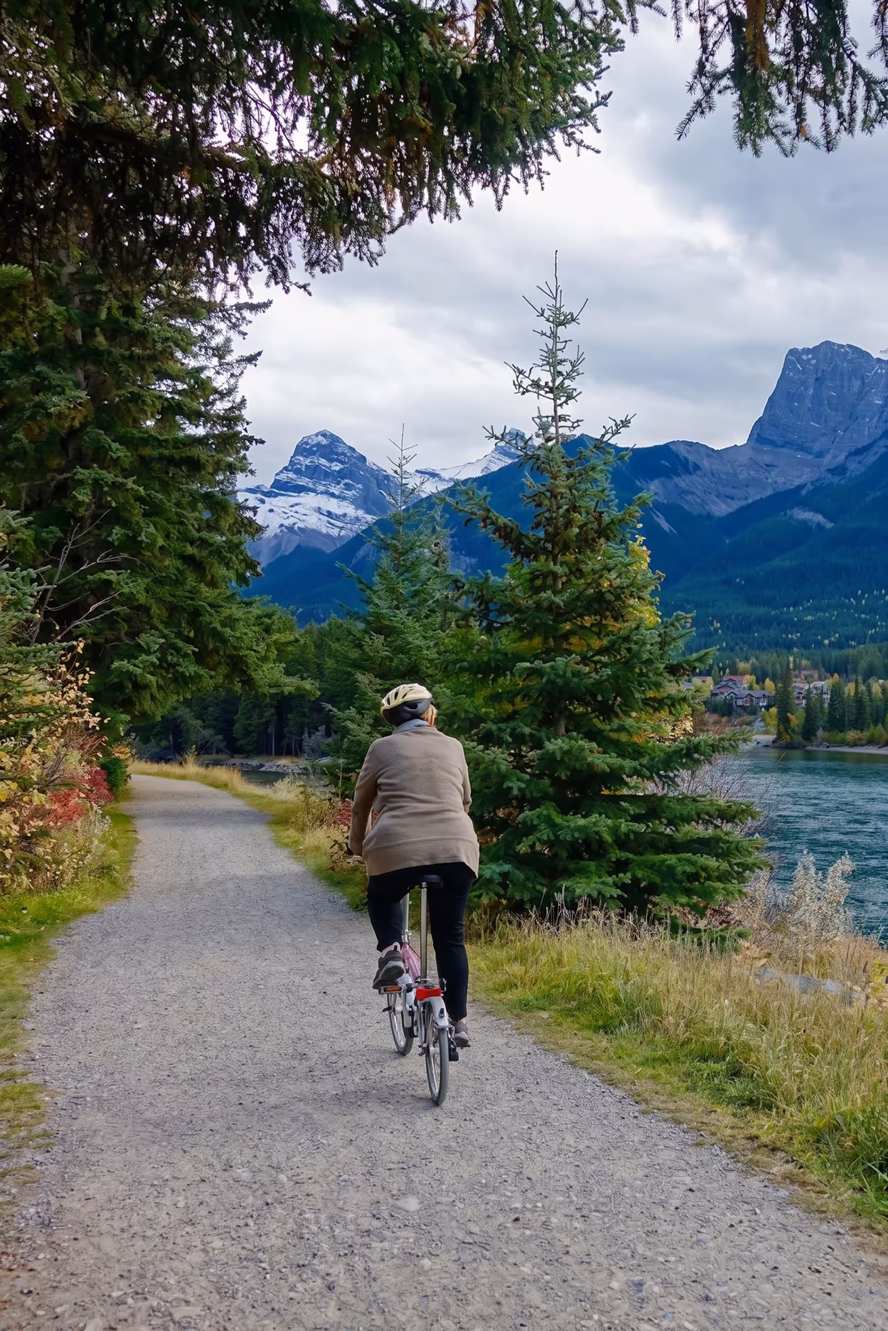 Person wearing a helmet riding a bike on a forest path with mountains and a lake in the background.