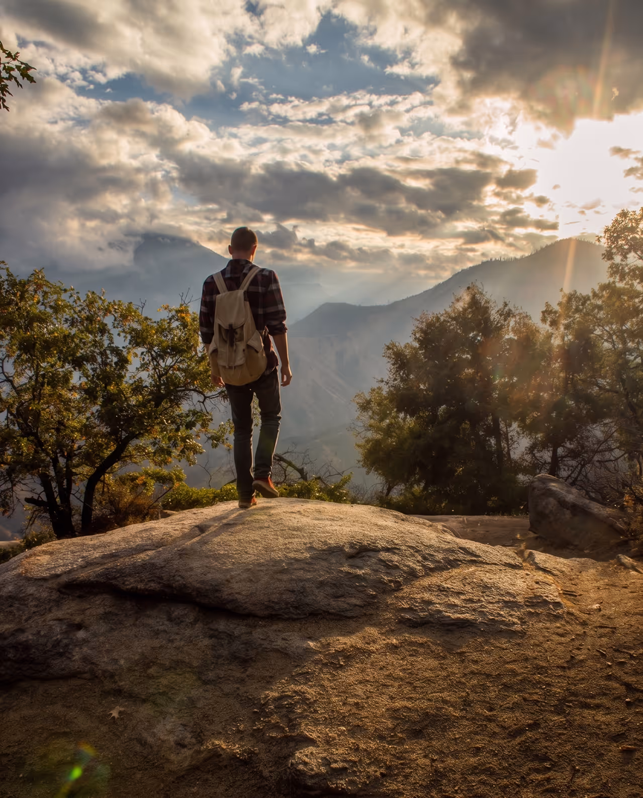 Hiker with a backpack standing on a rock overlooking a mountainous landscape under a cloudy sky at sunset.