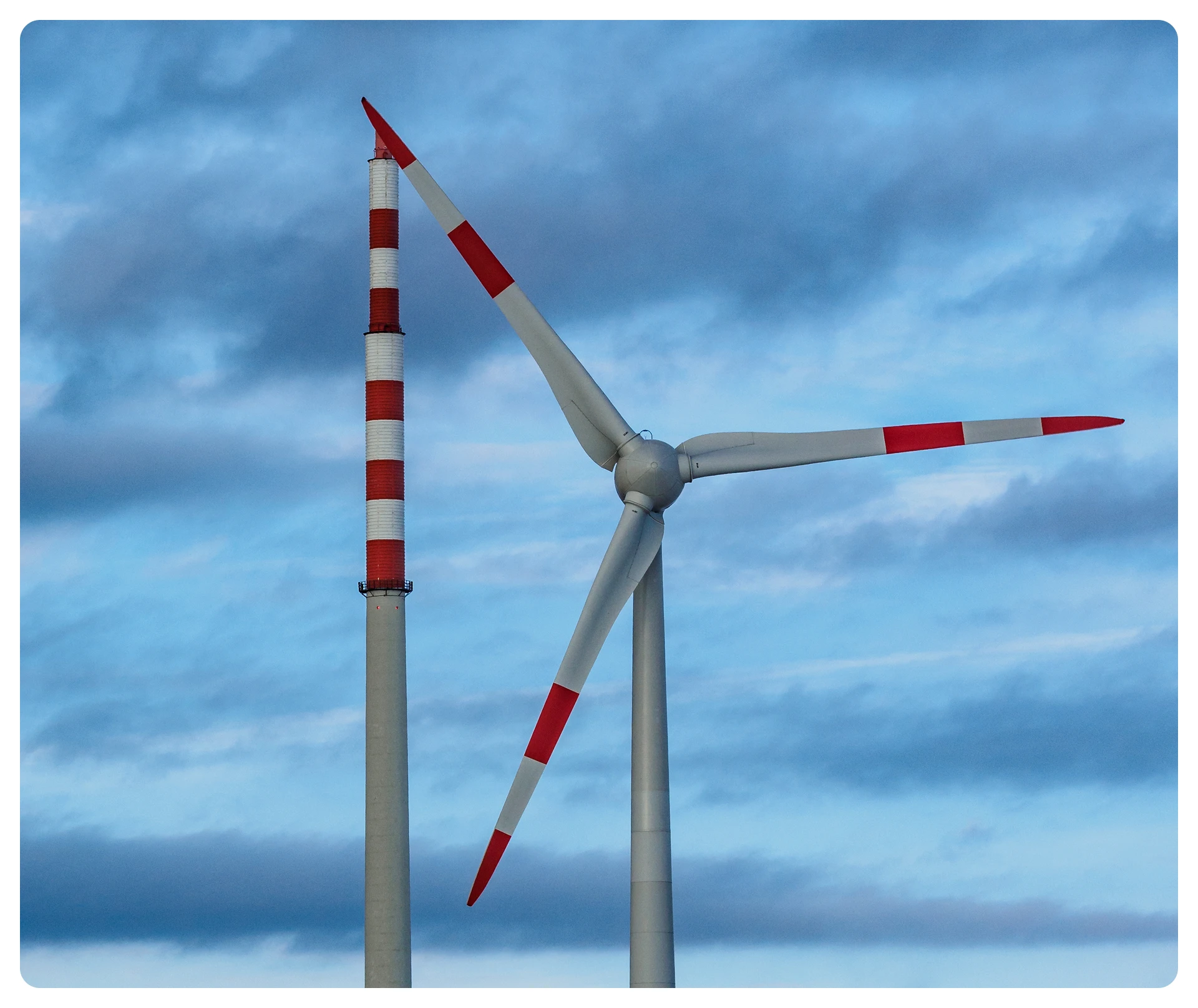 Close-up of a wind turbine and a tall red-and-white striped industrial chimney against a blue cloudy sky.