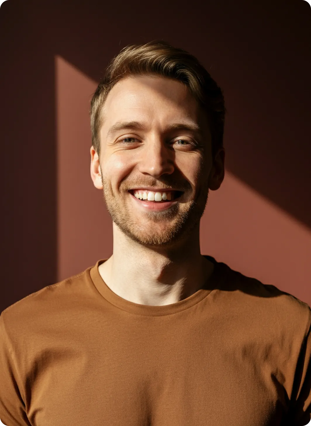 Smiling man with light brown hair and beard wearing a brown shirt against a dark reddish background.