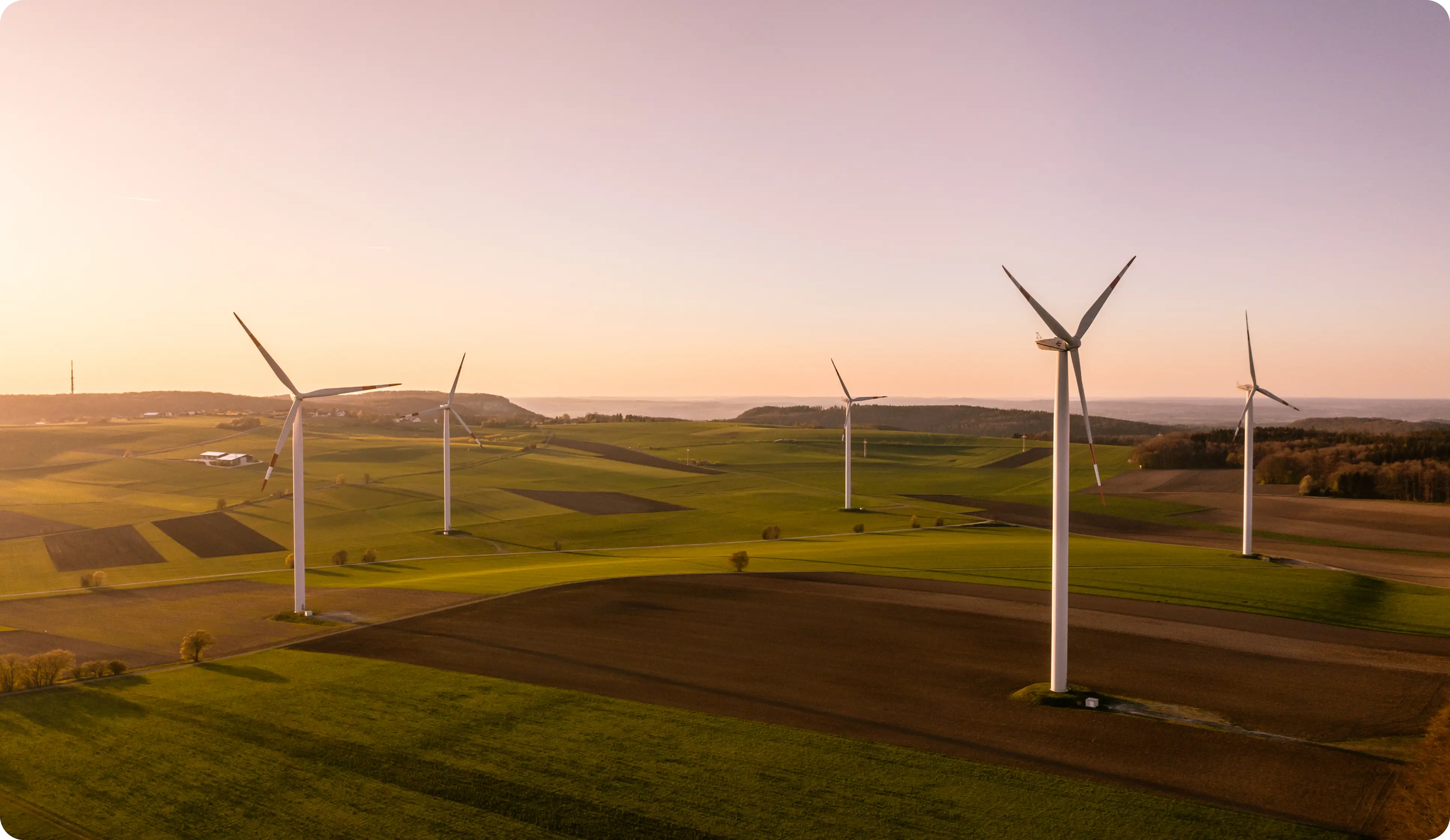 Wind turbines standing in a green and brown patchwork farmland under a clear sky at sunset.
