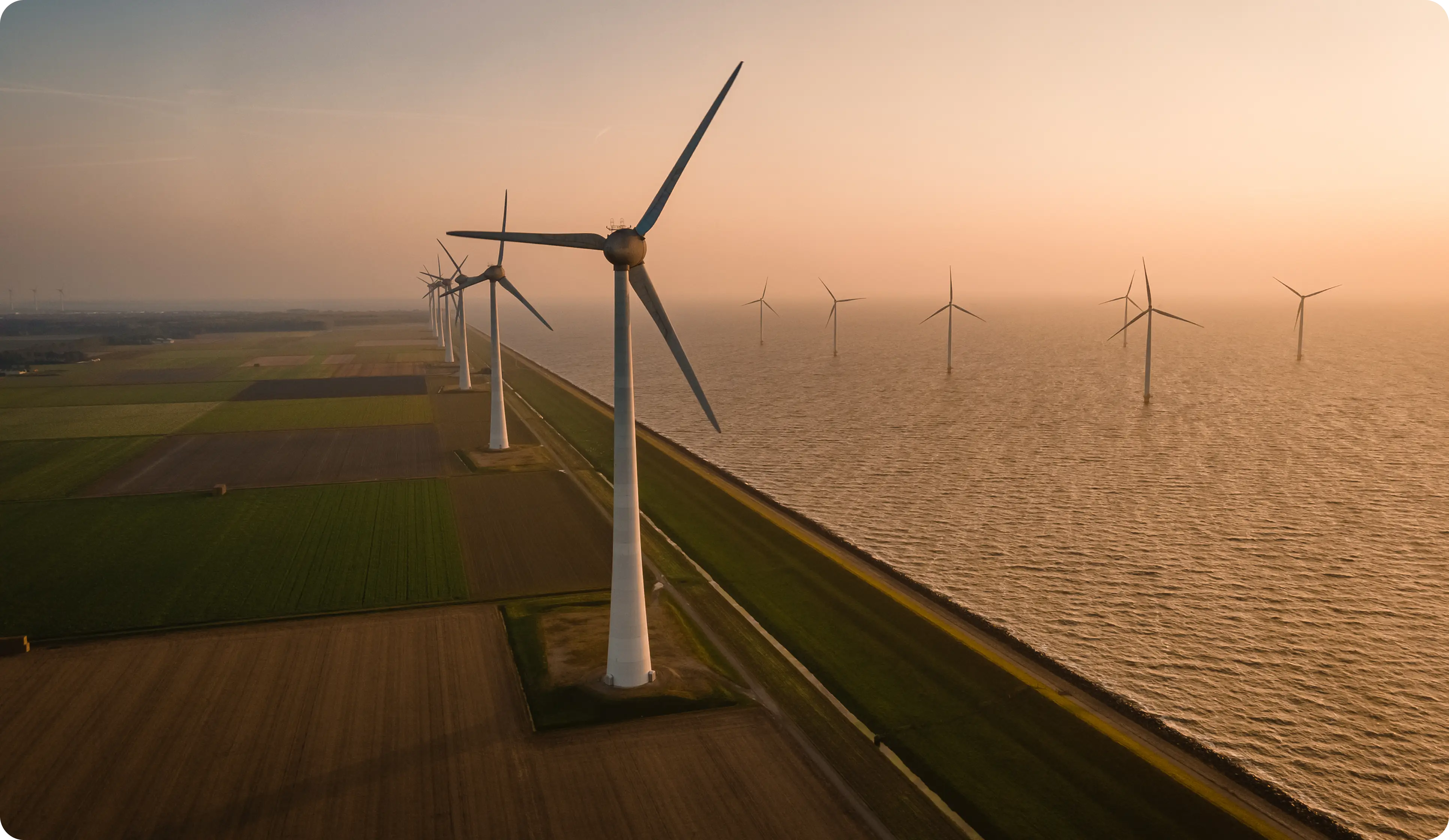 Aerial view of wind turbines along a coastline at sunset with fields on one side and water on the other.