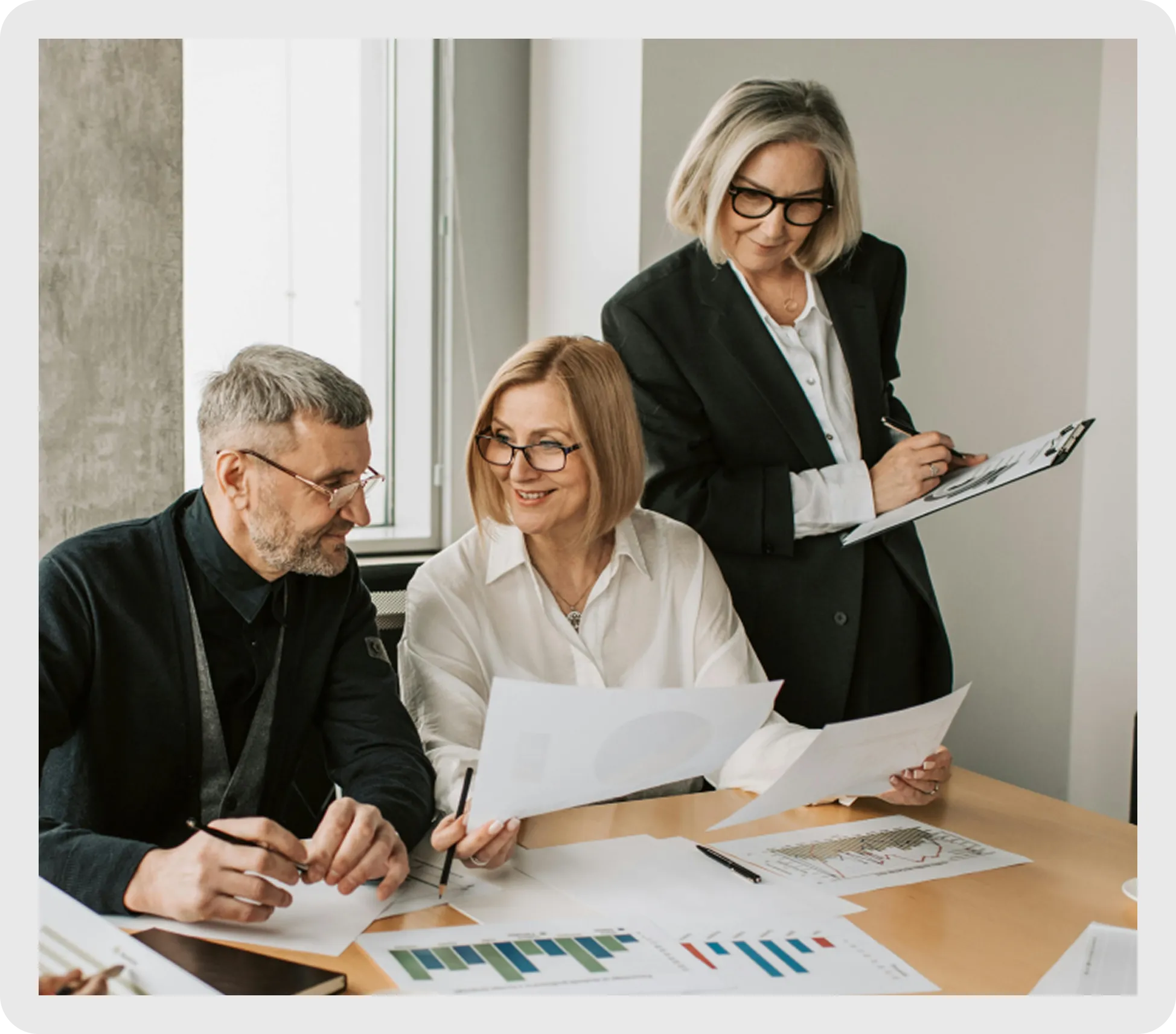 Three business professionals discussing charts and documents around a table in a bright office.