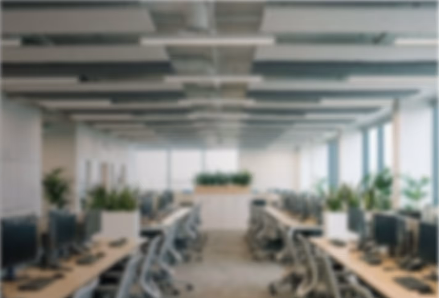 Blurred view of a modern office with rows of desks, chairs, computers, and green plants.