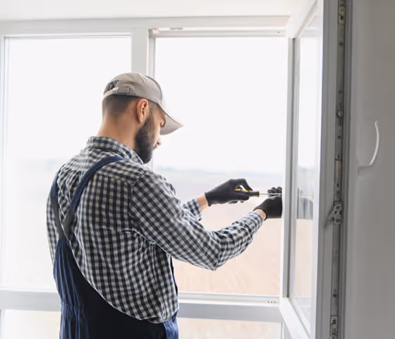 Man in checkered shirt and cap installing or measuring a white window frame while wearing black gloves.