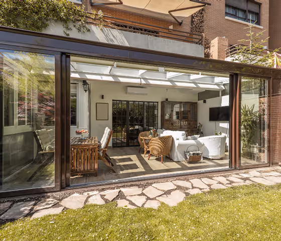 Sunlit patio with glass sliding doors, outdoor wicker furniture, dining table, and greenery.