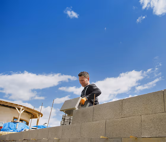 Construction worker wearing gloves placing a concrete block on a wall under a blue sky with clouds.