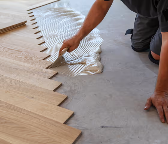 Person installing wooden flooring using adhesive and a notched trowel on a concrete surface.