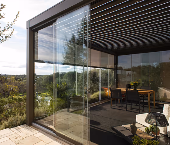 Modern outdoor patio with glass walls and a slatted roof overlooking a green garden, featuring a wooden dining table with black chairs and a sofa with decorative items.