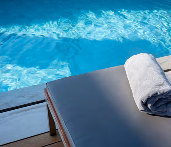 Rolled white towel on a gray poolside lounge chair next to clear blue swimming pool water.