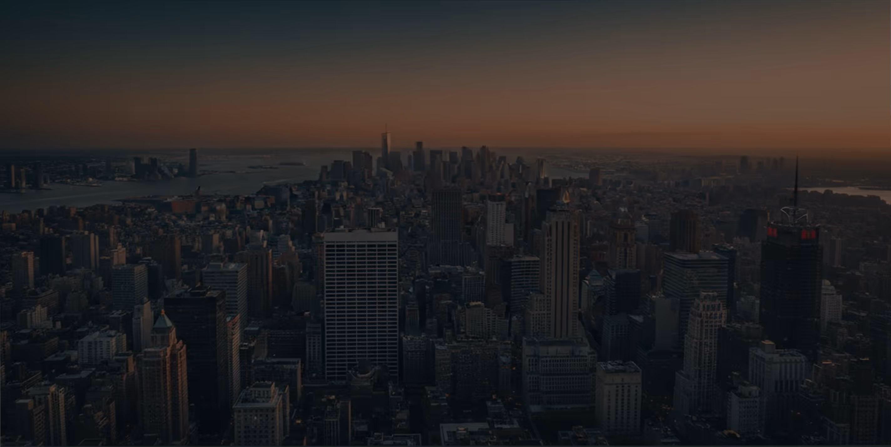 Cityscape of New York City at sunset with skyscrapers and a river in the background.