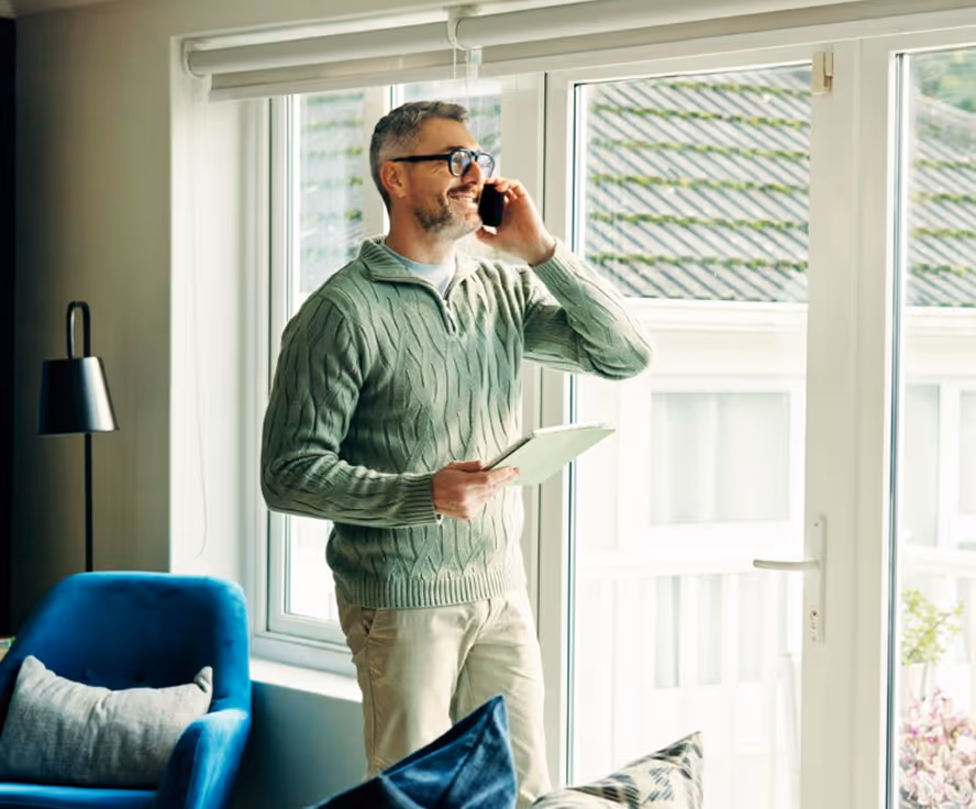 Man in glasses and green sweater holding a tablet and talking on a phone near a bright window.