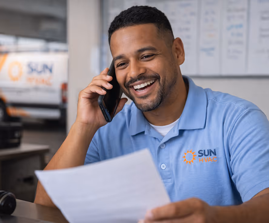 Smiling man in a blue Sun HVAC polo shirt talking on a phone while holding a document.