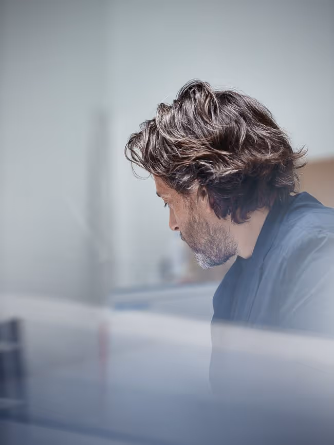 Man with wavy brown hair and beard, wearing a dark jacket, looking down in a softly blurred indoor setting.