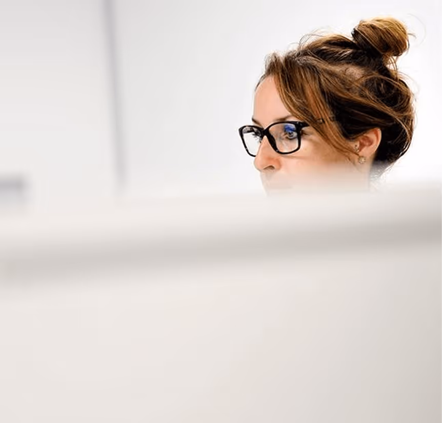 Woman with glasses and hair in a bun looking at a computer screen in a bright office.