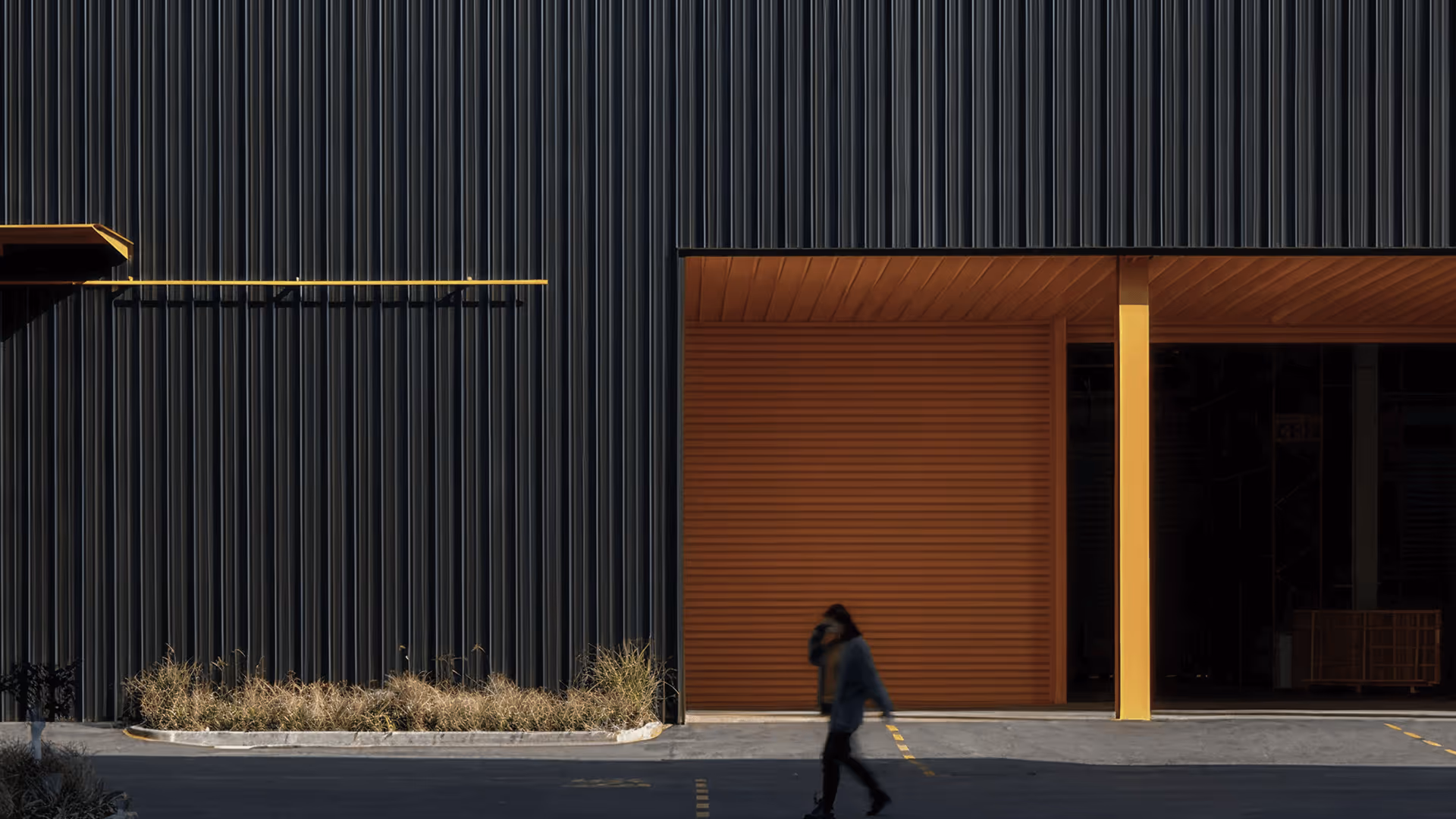 Person walking past a modern building with dark vertical panels and yellow architectural accents.