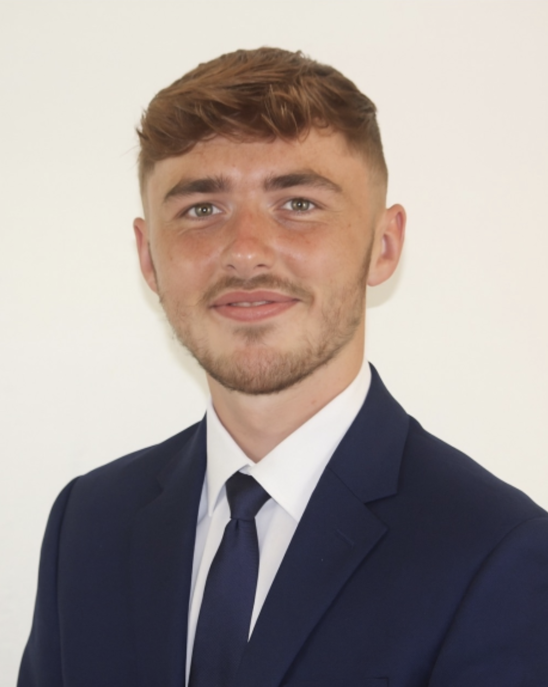 Young man with light brown hair and a short beard wearing a navy suit, white shirt, and navy tie smiling against a plain light background.