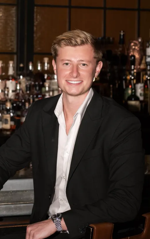 Smiling young man with blonde hair wearing a black suit jacket and white shirt seated in front of a bar with bottles in the background.