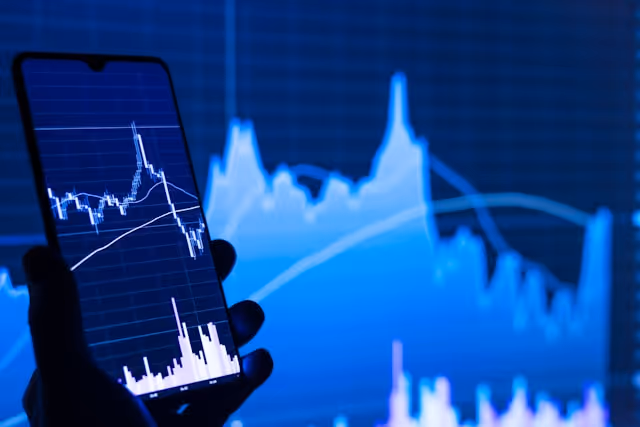 Hand holding smartphone displaying financial stock charts with blue background of similar charts.
