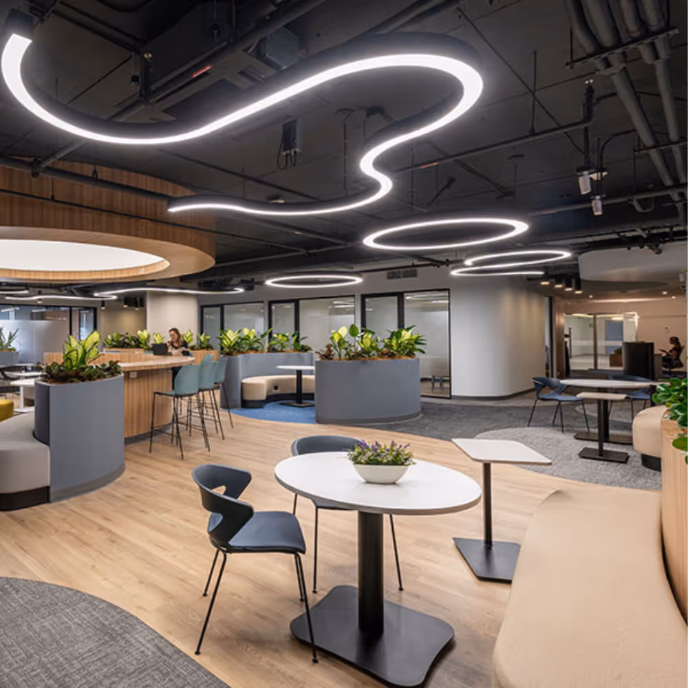 Modern office lounge area with circular and wavy ceiling lights, tables, chairs, and planted partitions.
