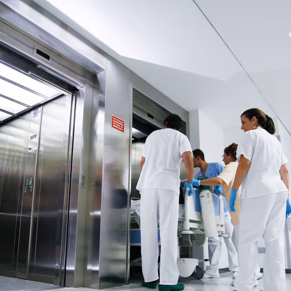 Medical staff in white scrubs and gloves pushing a patient on a hospital gurney into an elevator.
