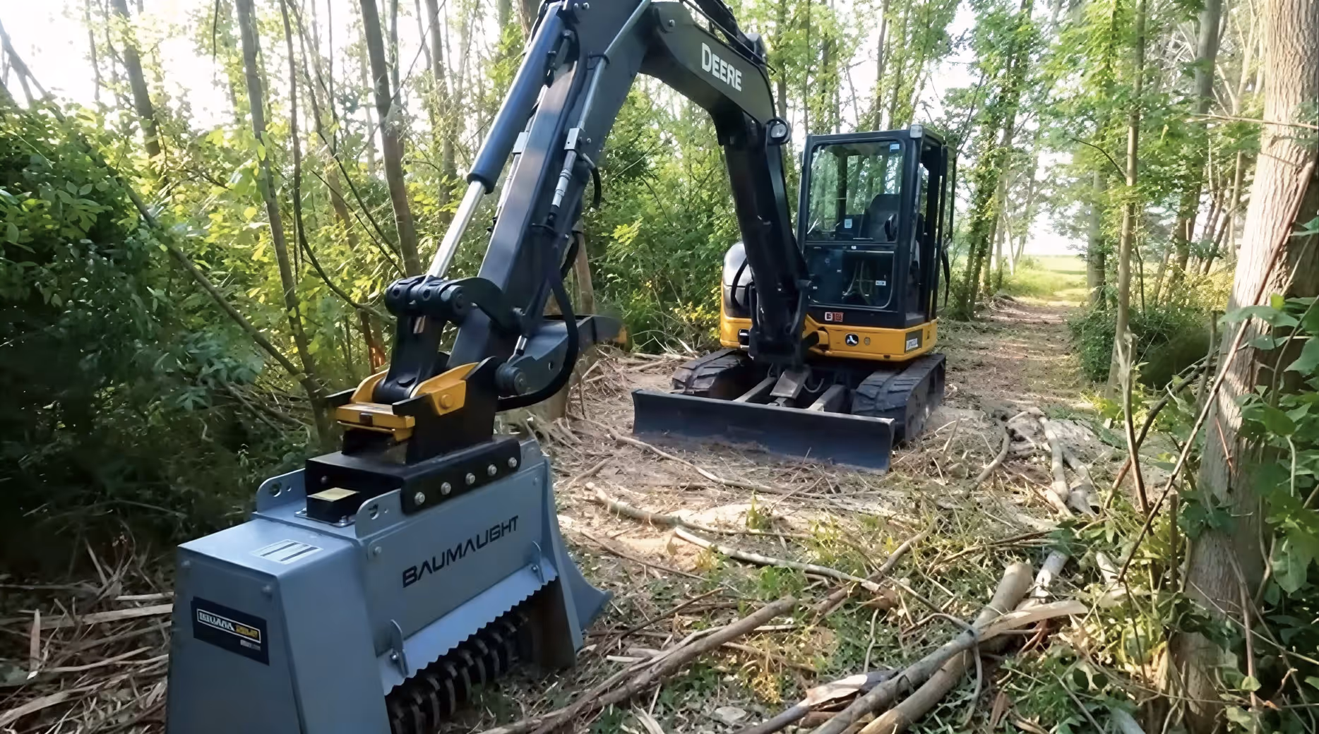 Yellow and black Deere mini excavator with a Baumalight mulcher attachment clearing branches in a wooded area.
