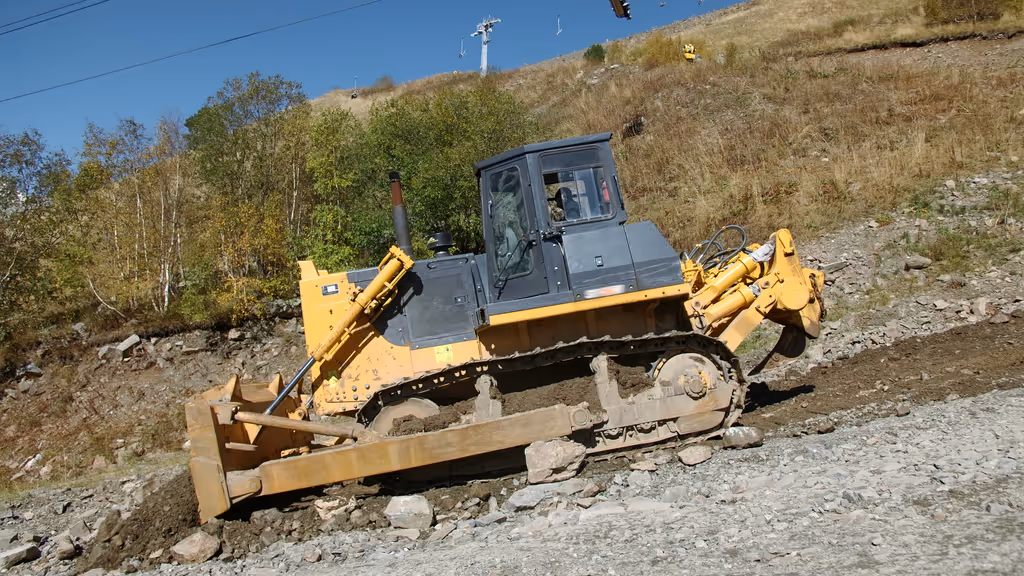 Yellow bulldozer with black cabin pushing dirt and rocks on a rough terrain near dry grass and trees under a clear blue sky.