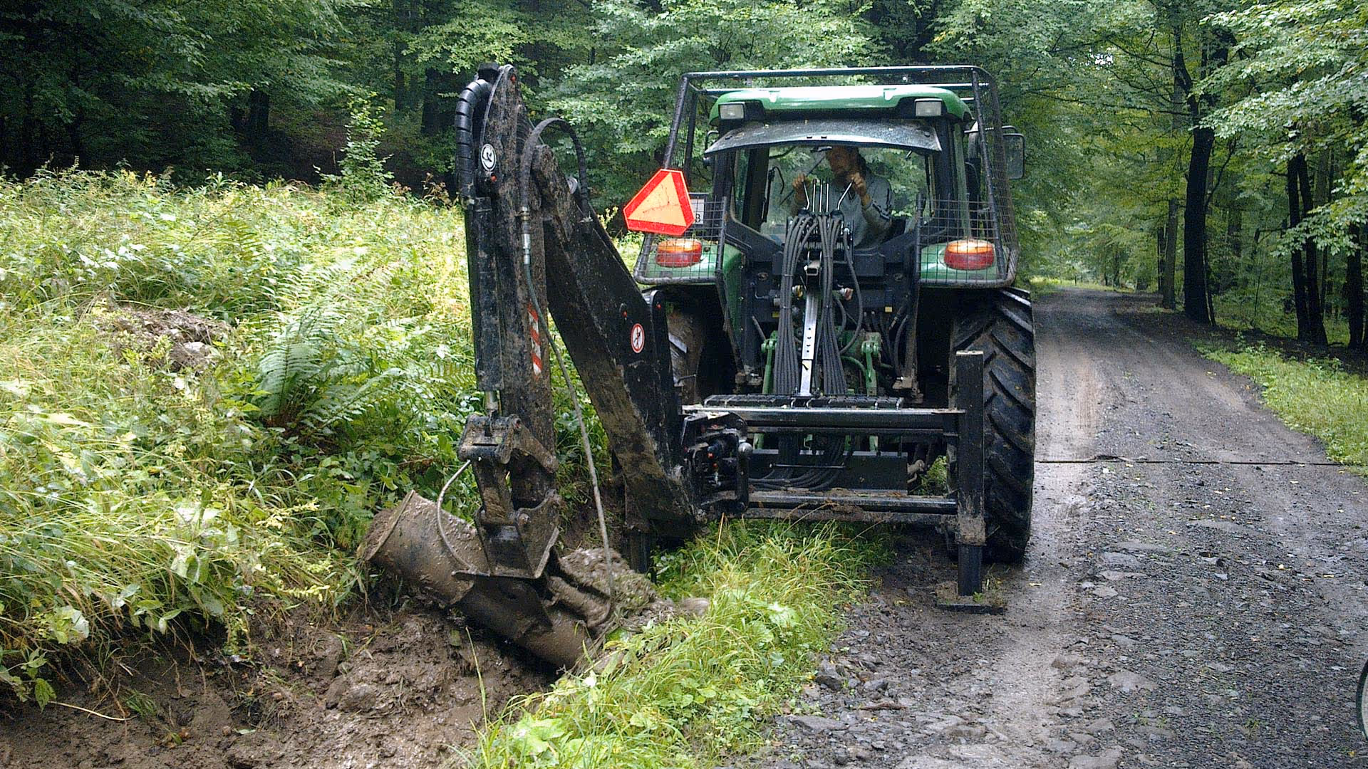 Tractor equipped with a backhoe digging a muddy ditch beside a forest road.