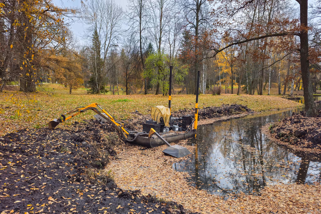 Small excavator dredging a narrow creek surrounded by autumn trees and fallen leaves.