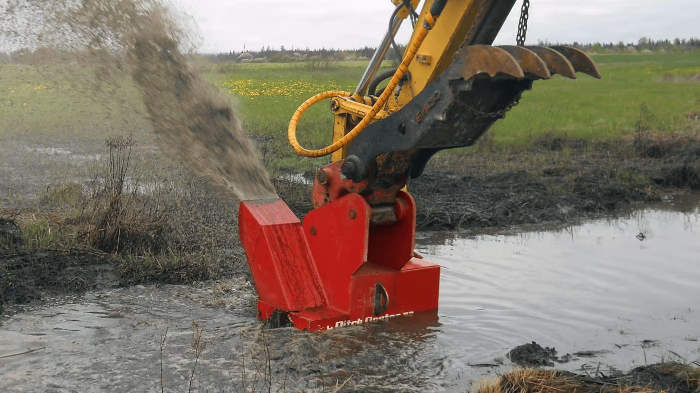 Excavator with a red attachment dredging and spraying muddy water in a wet grassy field.