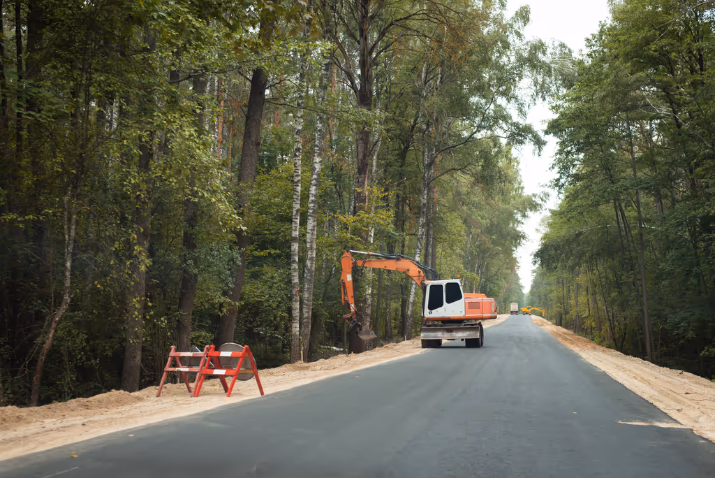 Orange excavator parked on a newly paved road surrounded by dense green forest.