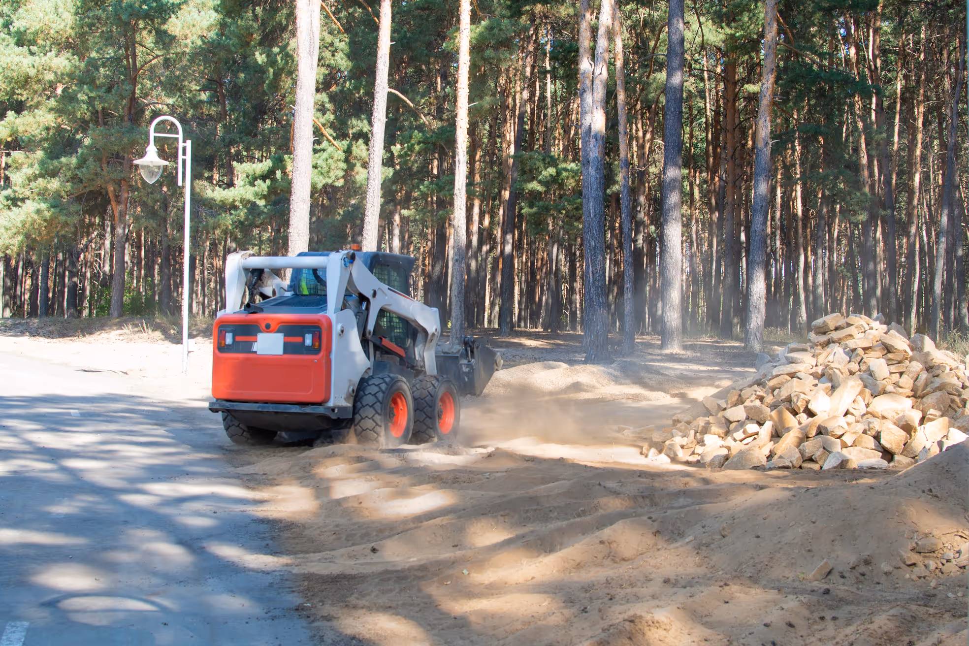 Small bulldozer moving dirt near a pile of rocks beside a forested area and a road lamp post.