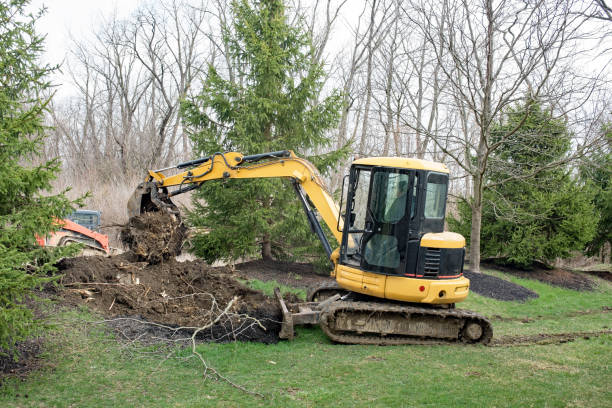 Yellow excavator digging and lifting soil in a grassy area surrounded by trees.