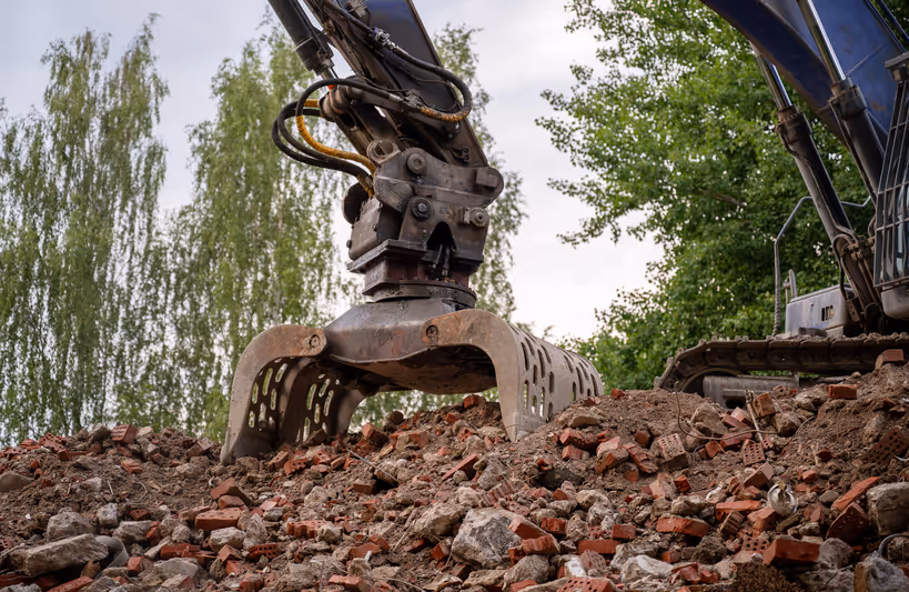 Heavy excavator claw gripping rubble and broken bricks at a demolition site with trees in the background.