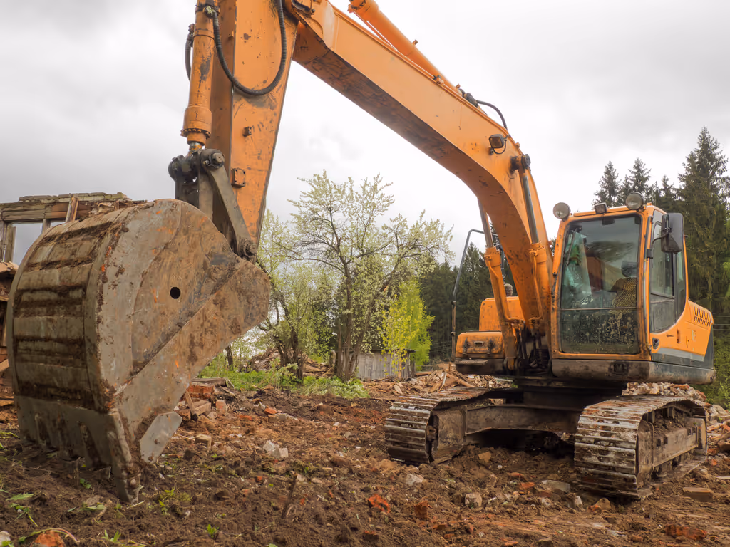 Orange excavator with a large bucket on a muddy demolition site with debris and trees in the background.