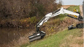 Excavator with a long arm digging or clearing near a creek surrounded by grass and trees.