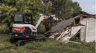 Bobcat excavator demolishing the side wall of a small wooden building surrounded by grass and trees.
