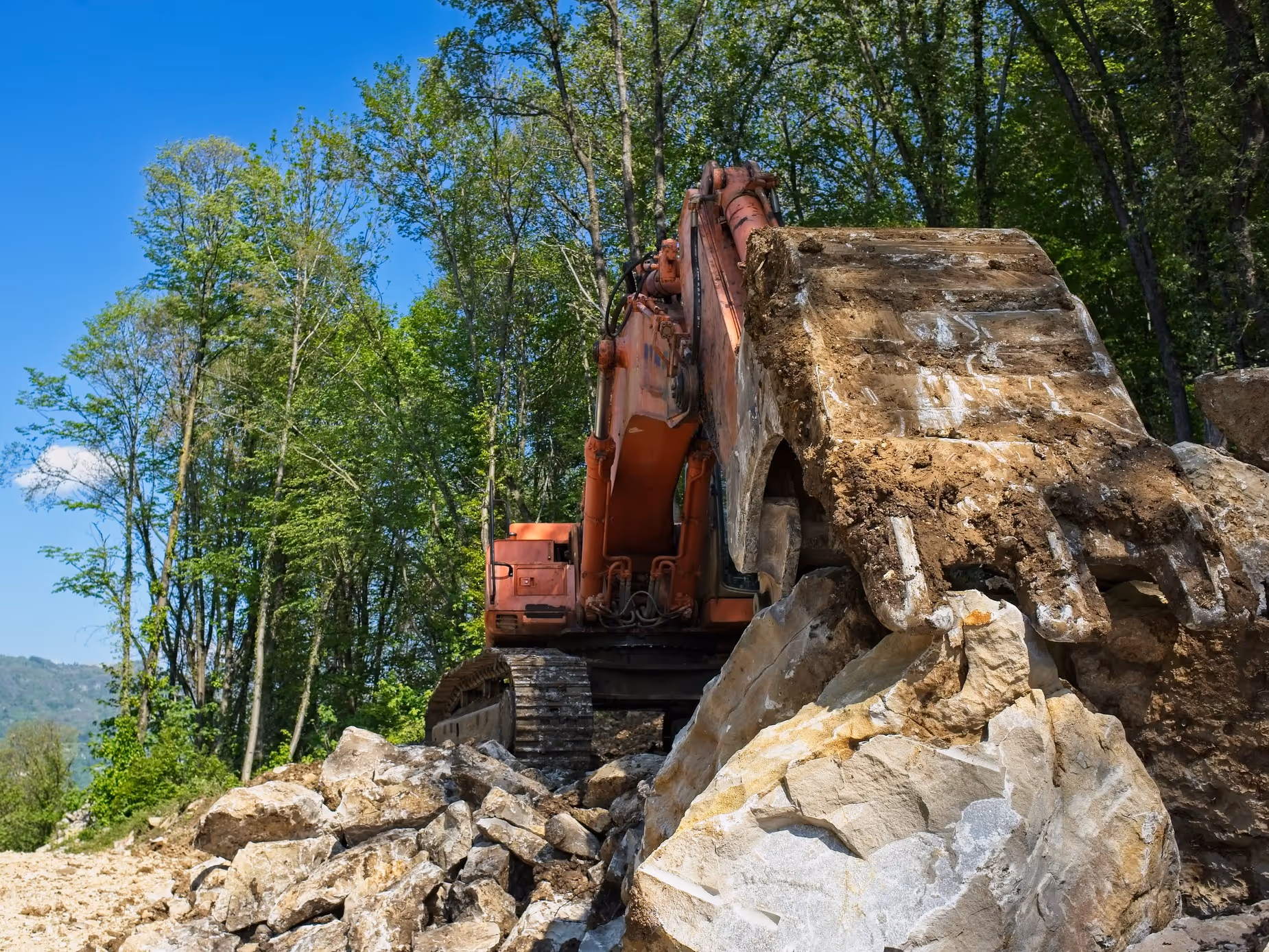 Large excavator bucket filled with dirt and rocks in a forested area under a blue sky.