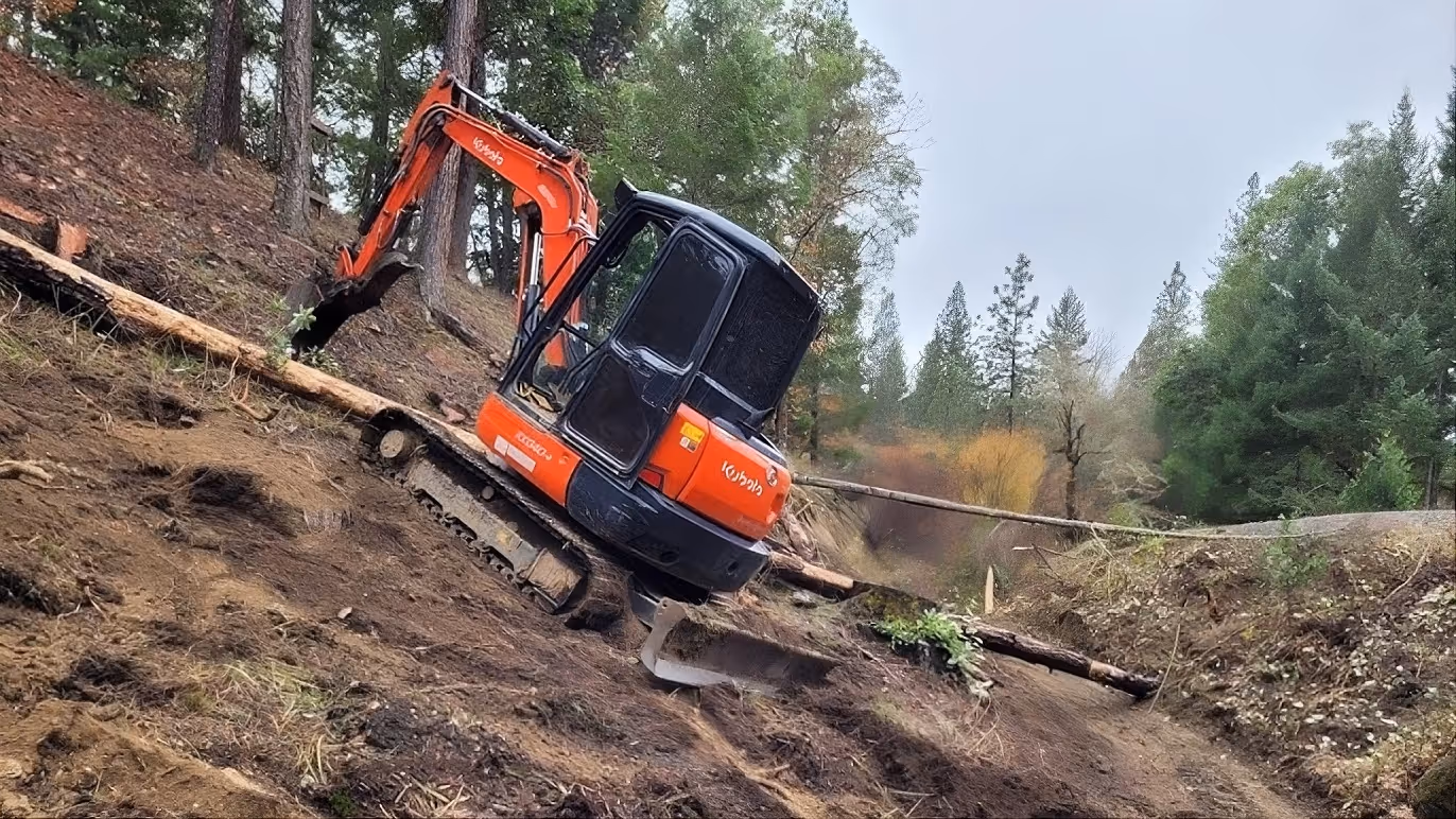 Orange Kubota excavator working on a steep, dirt-covered hillside surrounded by trees.