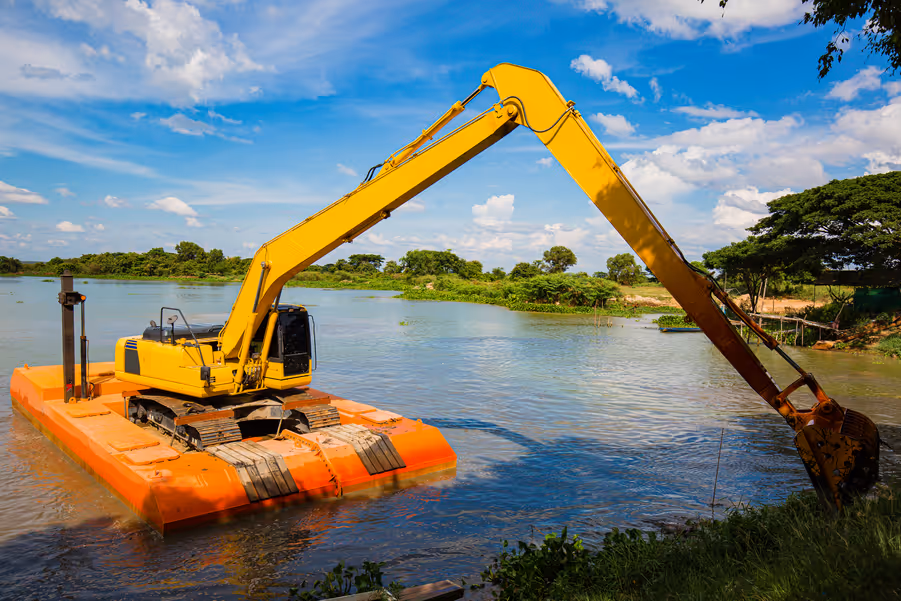 Yellow excavator mounted on an orange floating platform working in a river under a blue sky with scattered clouds.