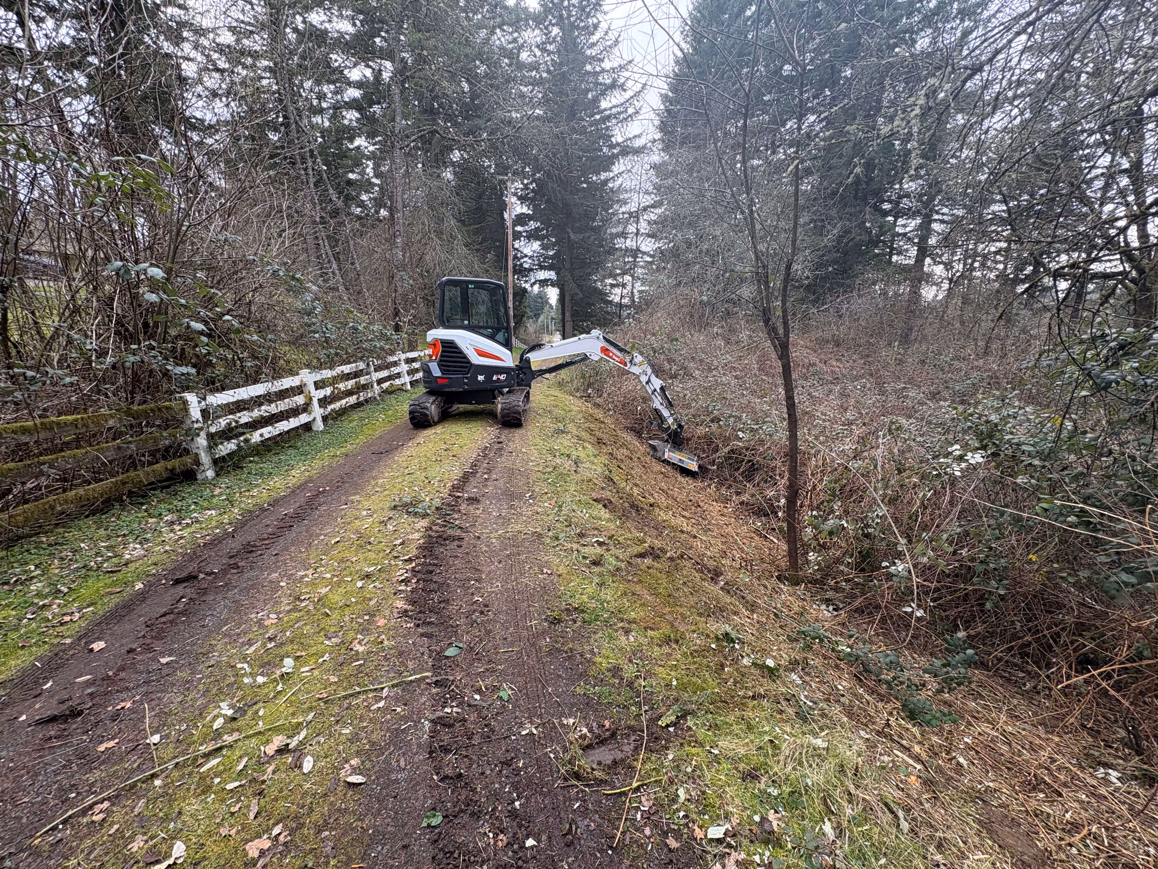 Excavator clearing brush and vegetation from a roadside ditch along a dirt path with trees and a white wooden fence.