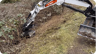 Excavator clearing brush and dirt from a ditch beside a grassy area.