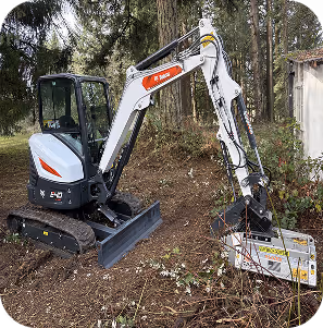 Compact white Bobcat excavator clearing brush in a wooded area near a small building.