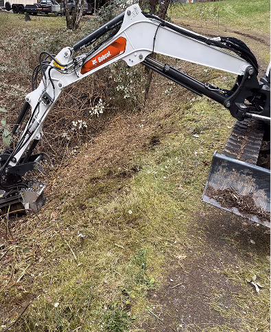 Bobcat excavator digging on a grassy hillside with brush and a dirt path nearby.