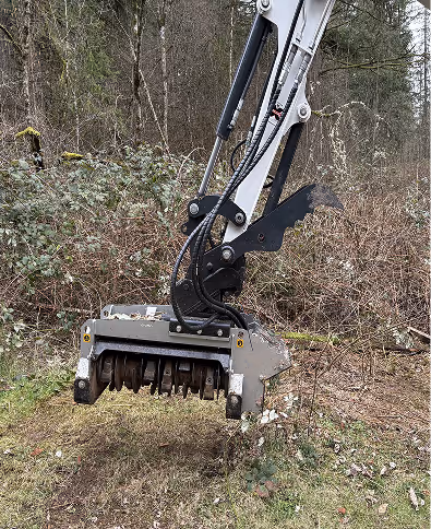 Close-up of an excavator mulcher attachment above ground with forest brush in the background.