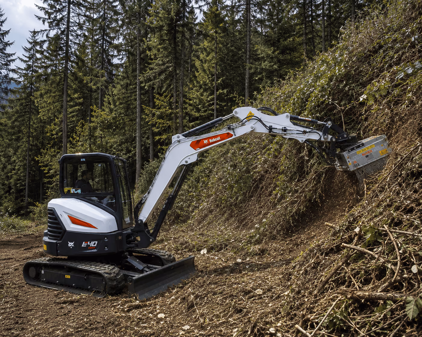 Small Bobcat excavator clearing brush on a forest hillside with trees in the background.