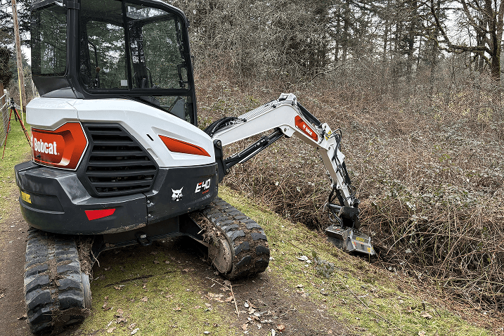 Bobcat E40 mini excavator clearing brush and dirt from a ditch near a wooded area.