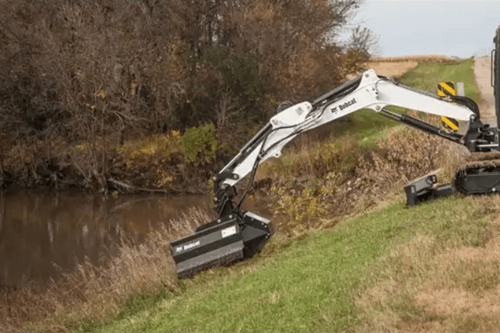 Bobcat excavator clearing vegetation on a grassy slope beside a creek or pond.