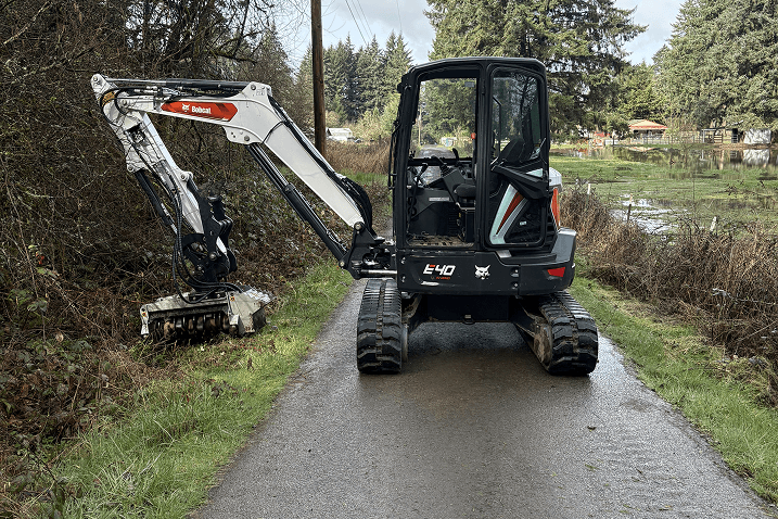 Black and white Bobcat E40 excavator clearing brush on a narrow wet road in a rural area.